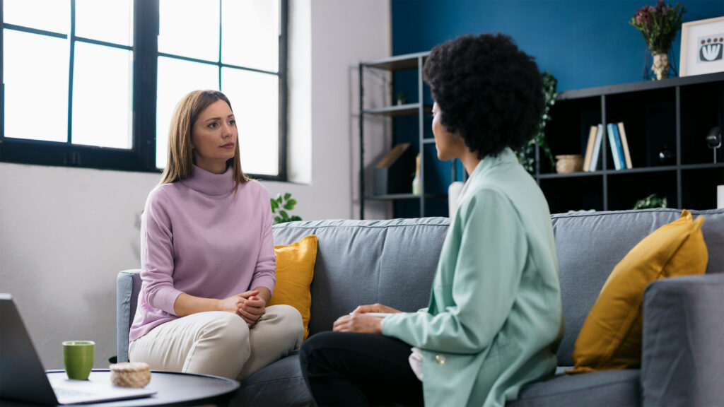 Two women sitting on a couch engaged in a serious conversation in a modern living room setting.
