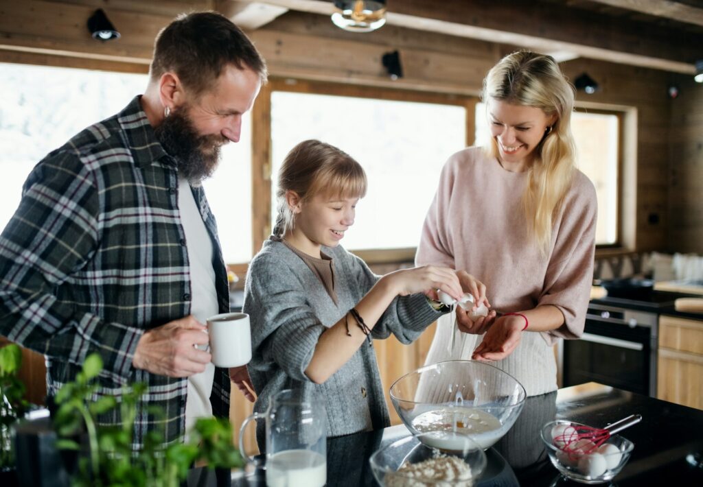 Family enjoying time together while baking in a cozy kitchen, with parents smiling as their child cracks eggs into a bowl.