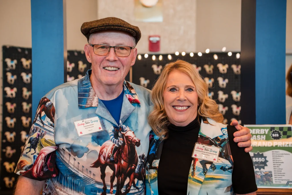 DCCH photo-11.webp | Two smiling attendees in matching horse-racing themed shirts pose together near Derby Bash raffle signage.