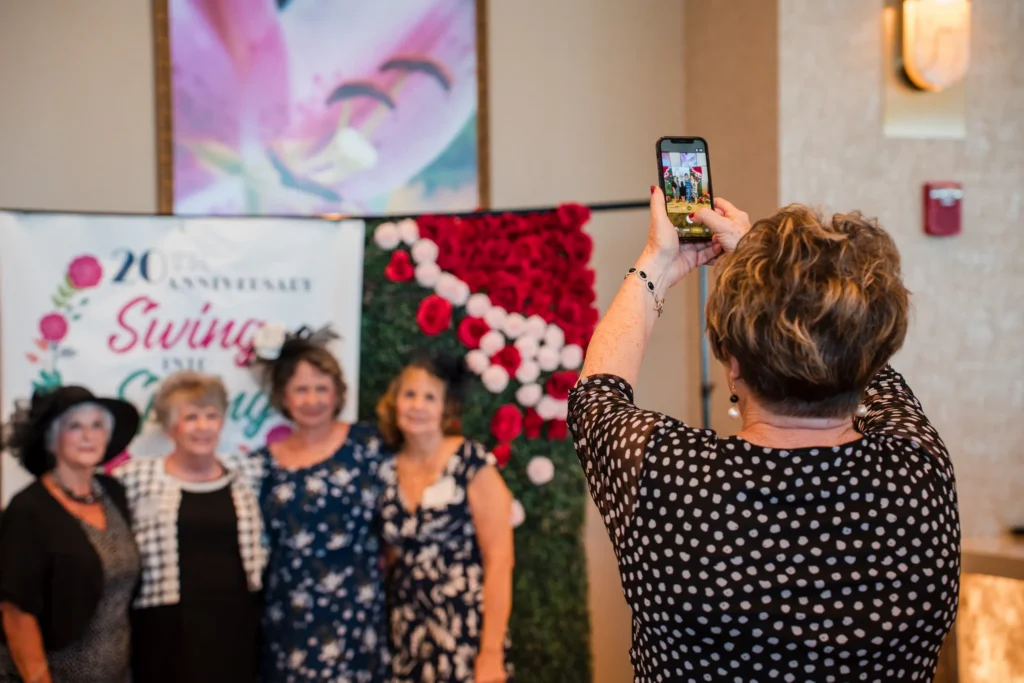 DCCH photo-12.webp | Guest taking a phone photo of attendees posing in front of the 20th Anniversary Swing Into Spring backdrop.